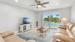 A spacious living room with a ceiling fan, a flat-screen TV, a wooden coffee table, and a beige leather sofa. - ⛱️ Beach Front Condo🚀🌴👙🚢 (Cocoa Beach)