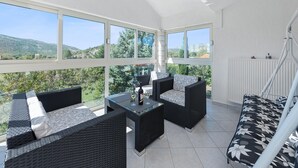 A sunlit patio with wicker furniture, a glass table, and a view of green hills. - Beautiful holiday home with private sauna (Neoric)