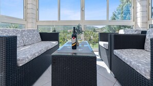 A sunlit patio with wicker furniture, a table with a bottle and glass, and a scenic view through large windows. - Beautiful holiday home with private sauna (Neoric)