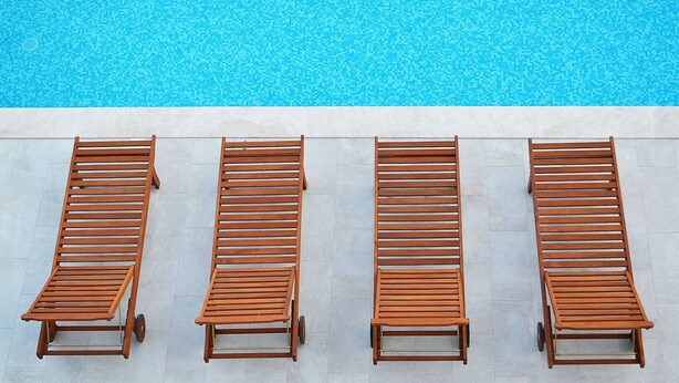 A row of wooden lounge chairs aligned parallel to a swimming pool.