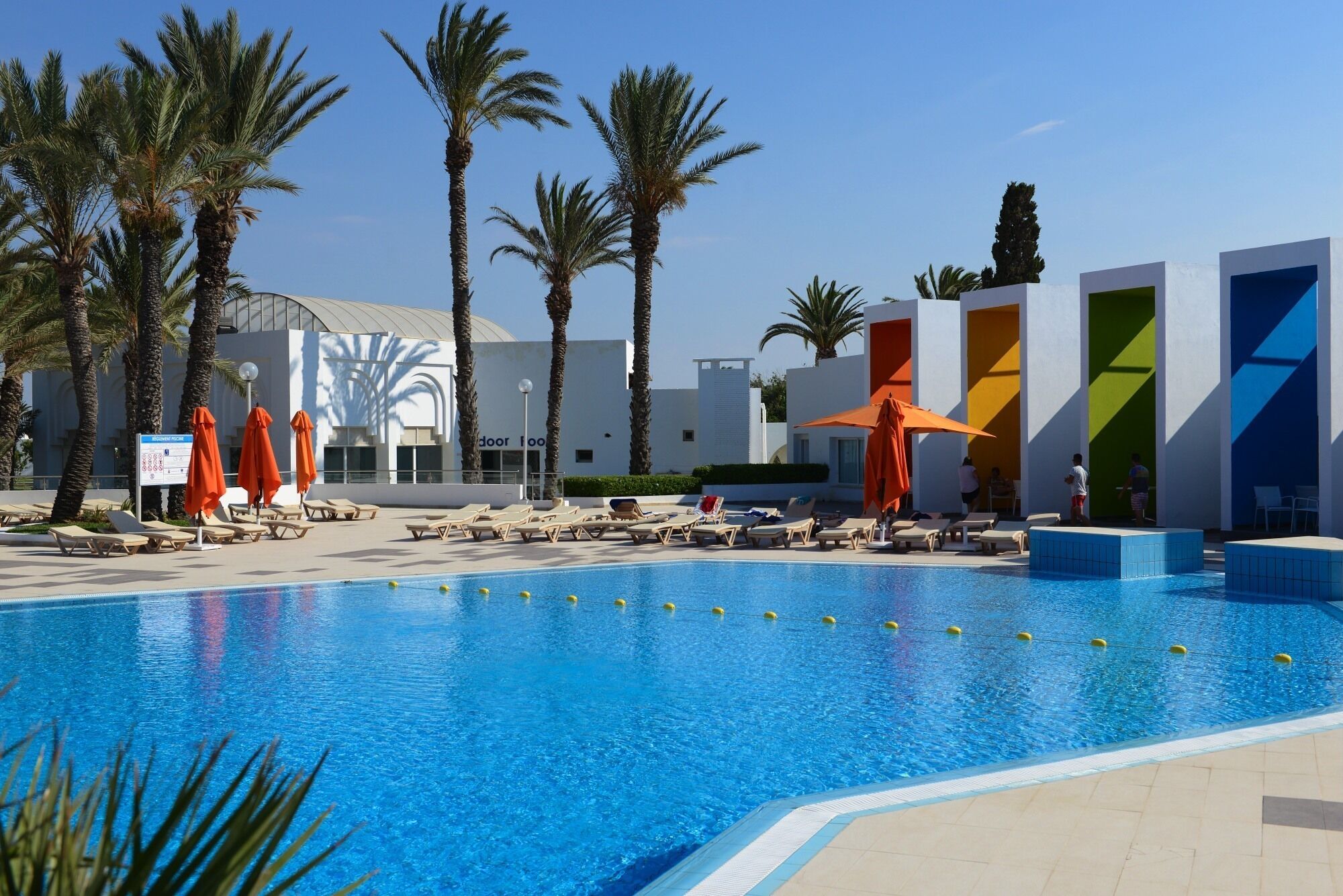Ein Swimmingpool mit Liegestühlen und Sonnenschirmen, umgeben von Palmen und modernen Gebäuden.