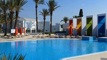 Ein Swimmingpool mit Liegestühlen und Sonnenschirmen, umgeben von Palmen und modernen Gebäuden.