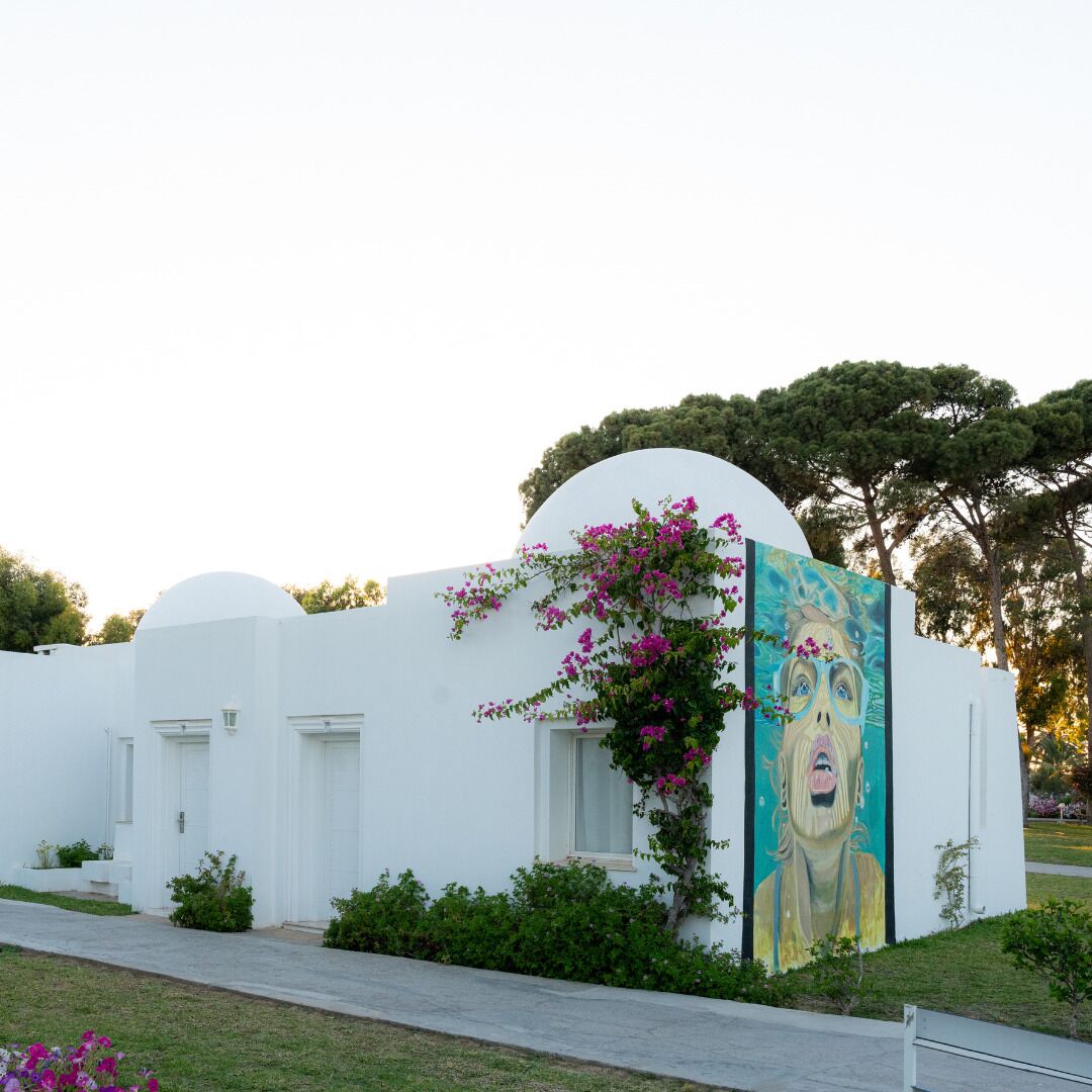 Un edificio blanco con un mural lateral, rodeado de vegetación y árboles.