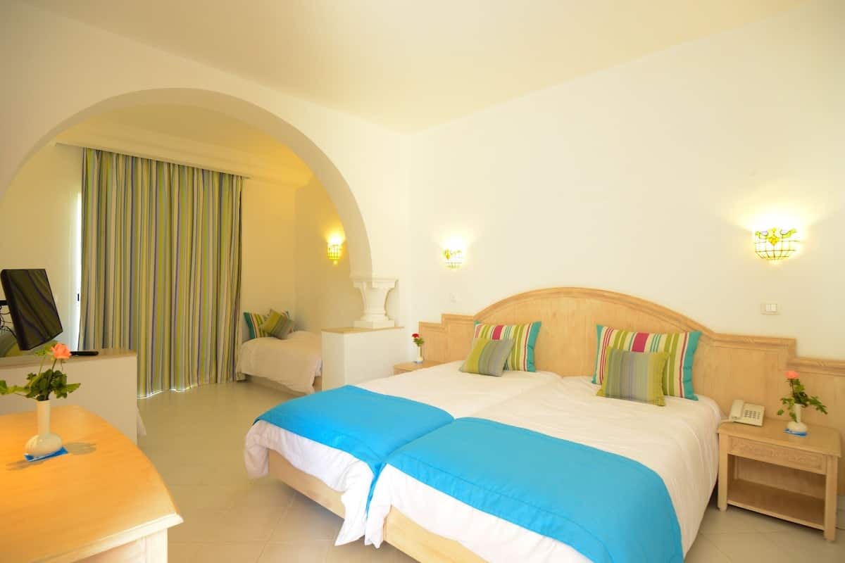 Ein Hotelzimmer mit einem großen Bett, zwei Nachttischen, einem Schreibtisch mit Computer und einem großen, bogenförmigen Durchgang zu einem weiteren Raum.