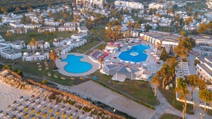 Eine Vogelperspektive auf ein Resort mit mehreren Gebäuden, einem großen zentralen Pool und einem Strandbereich mit Liegestühlen.