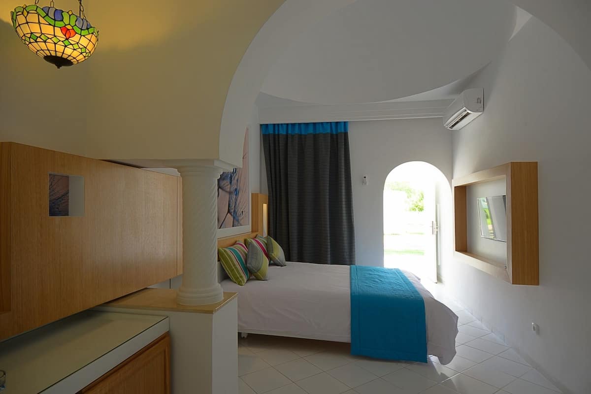 Ein Schlafzimmer mit einem Bett, einem Schreibtisch, einem Stuhl, einem Fenster mit Vorhängen und einer Deckenleuchte.