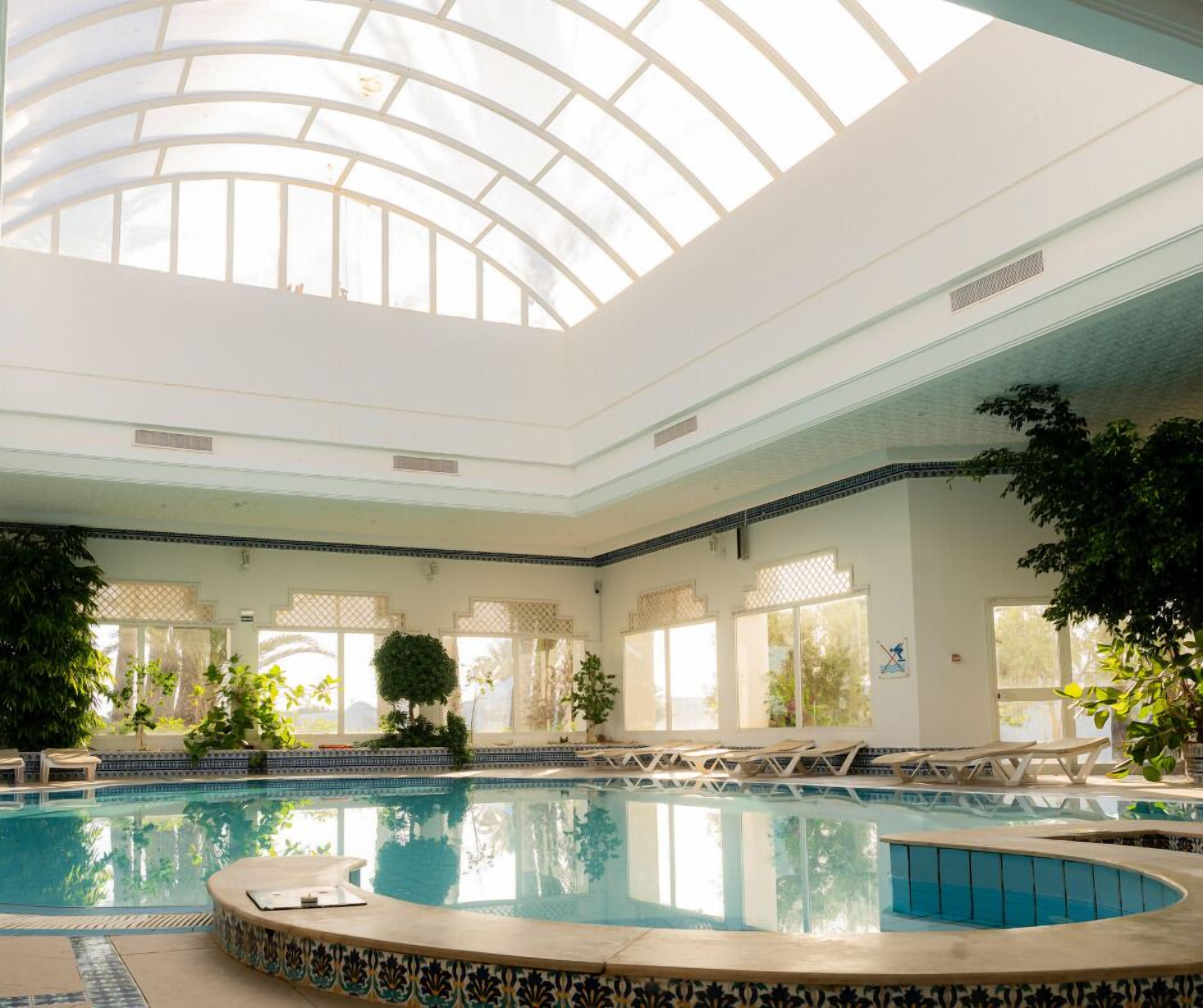 An indoor swimming pool with a glass ceiling, surrounded by lounge chairs and potted plants.