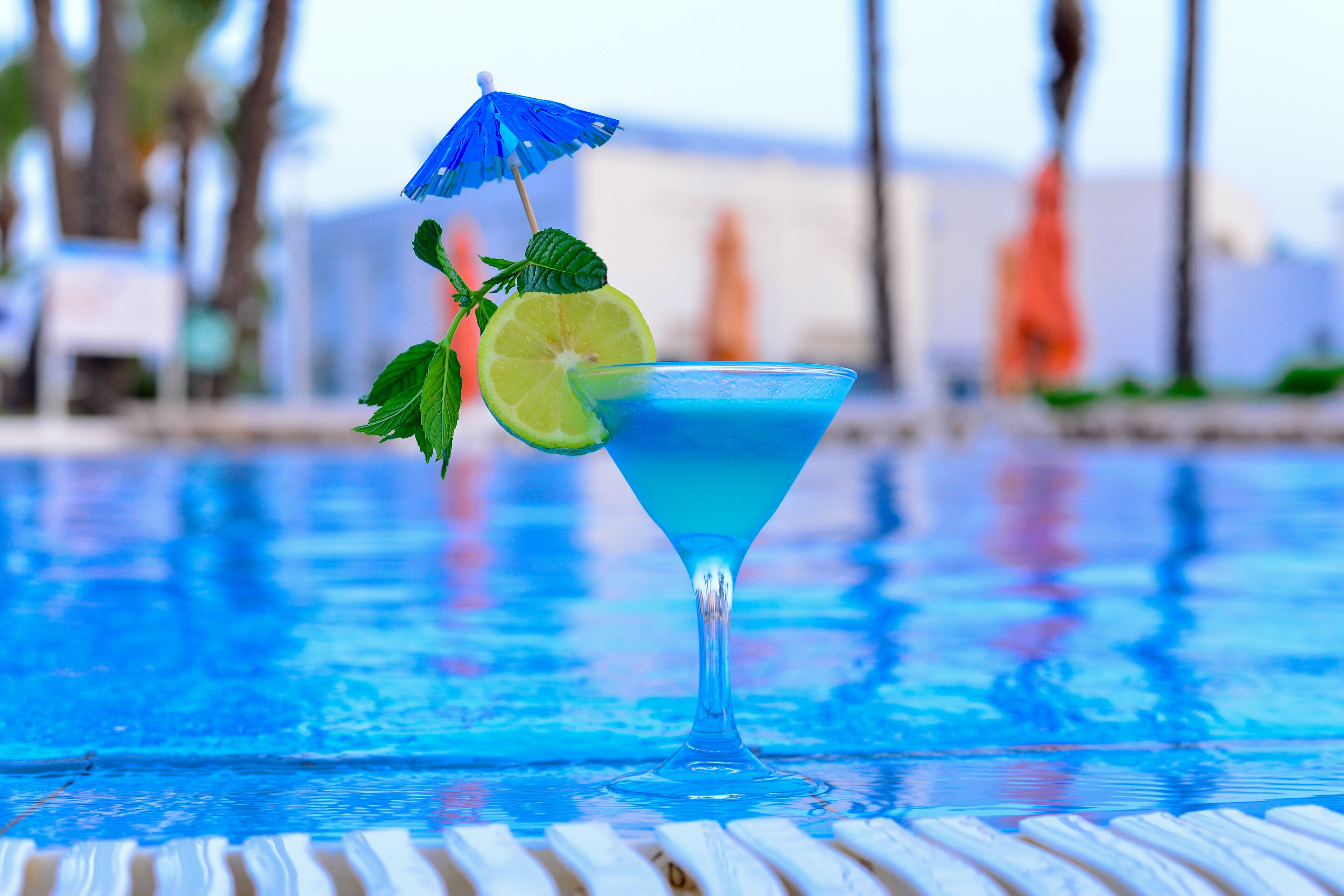 A blue cocktail with a lime slice and mint garnish on a poolside table.