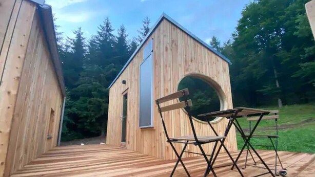 Une cabane en bois dotée d’une fenêtre ronde et d’une terrasse.