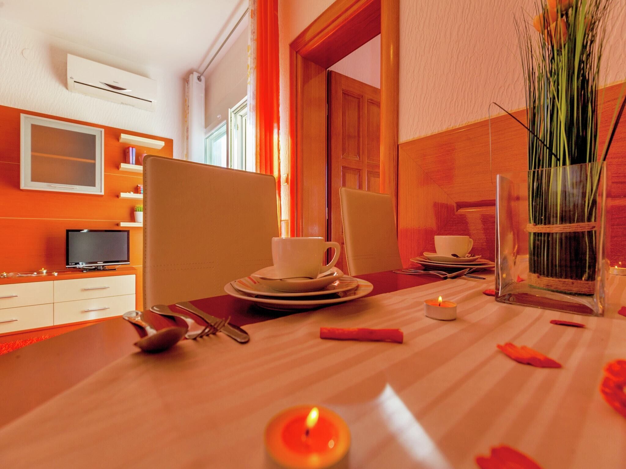 A dining area with a table set for two, including plates, cups, and cutlery. There is a TV on a stand to the left and a window with curtains.