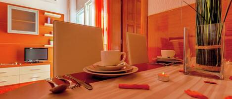 A dining area with a table set for two, including plates, cups, and cutlery. There is a TV on a stand to the left and a window with curtains.