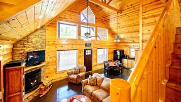 A cozy log cabin interior with a stone fireplace, leather sofas, a dining area, and a wooden staircase.