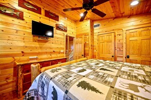Una habitación de cabaña de madera con una cama, un ventilador de techo, un televisor montado y puertas de madera.