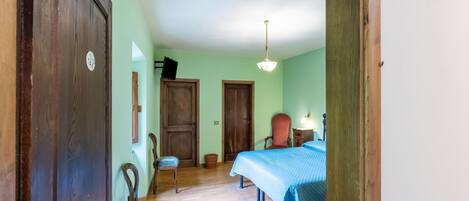 Ein Zimmer mit Holzboden, einem Bett mit blauer Bettdecke, einem Stuhl, einem Schreibtisch mit Lampe und einer Tür.