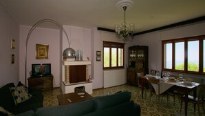 Una sala de estar con chimenea, un comedor con una mesa preparada para dos personas y una cocina con ventana que da a zonas verdes.