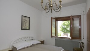 Un dormitorio con cama, ventana, radiador, silla, mesita de noche y una araña.
