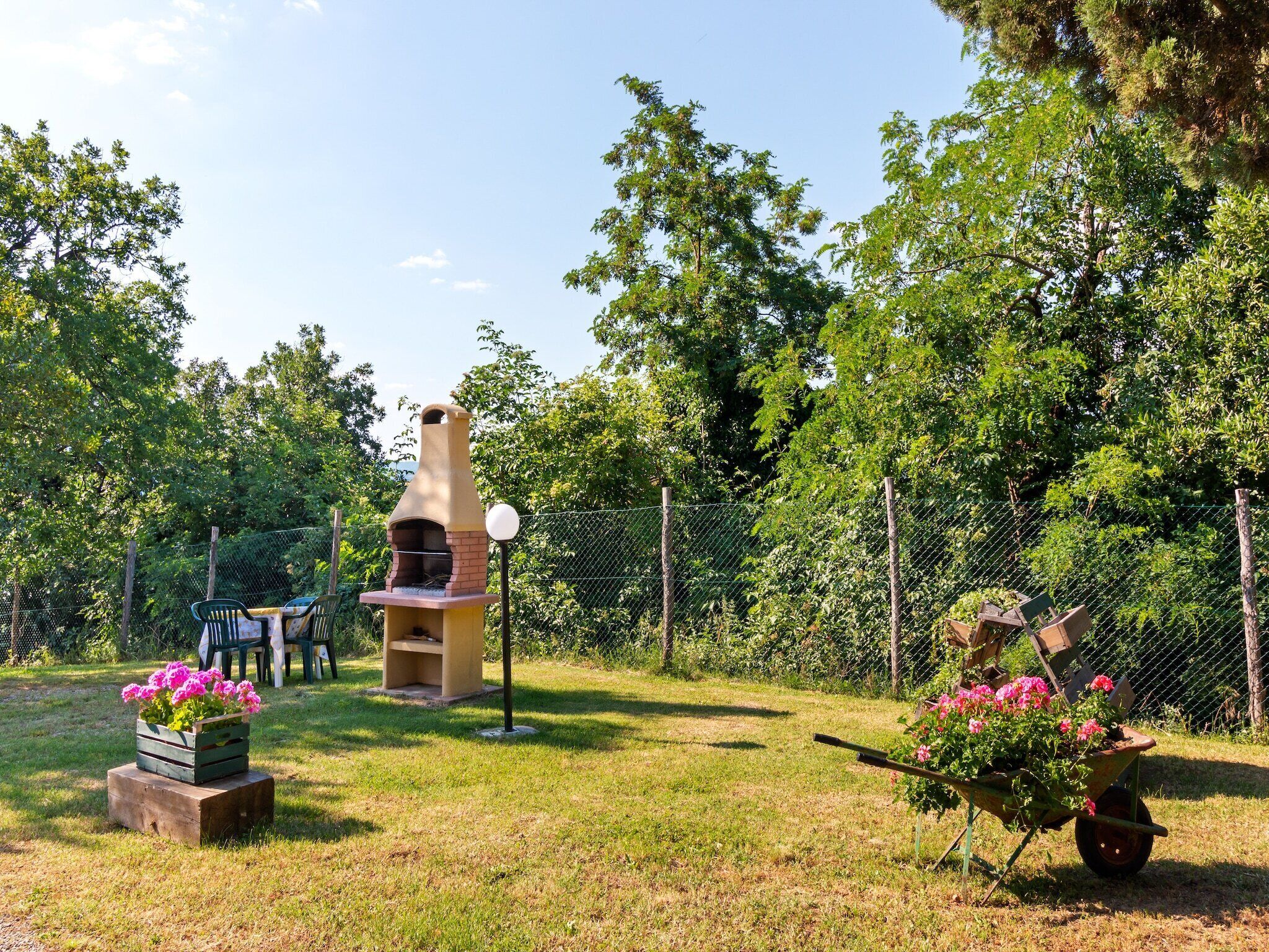 A backyard with a brick outdoor oven, a wooden table, and chairs. There are flower pots with pink flowers and a wheelbarrow with more flowers.