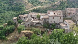 Un pueblo en una ladera con edificios de piedra, algunos con tejados de tejas, y un puente de piedra que conecta las construcciones.