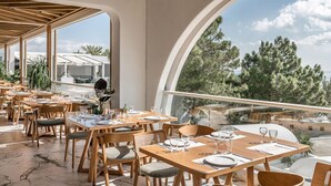 A restaurant with wooden tables and chairs, large windows, and a view of trees and mountains. - Kakkos Bay (Ierapetra)