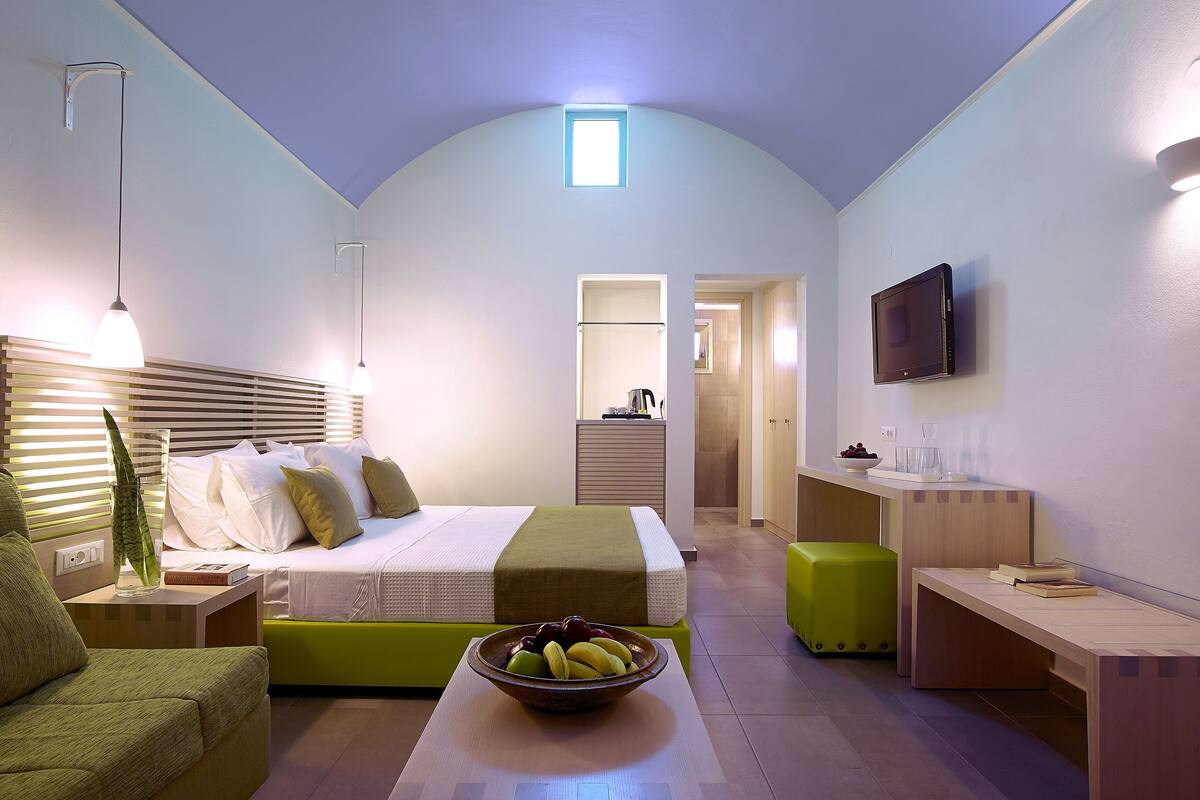 A modern hotel room with a bed, bedside table, green ottoman, a bowl of fruit, a desk, a flat-screen TV, and a small window.