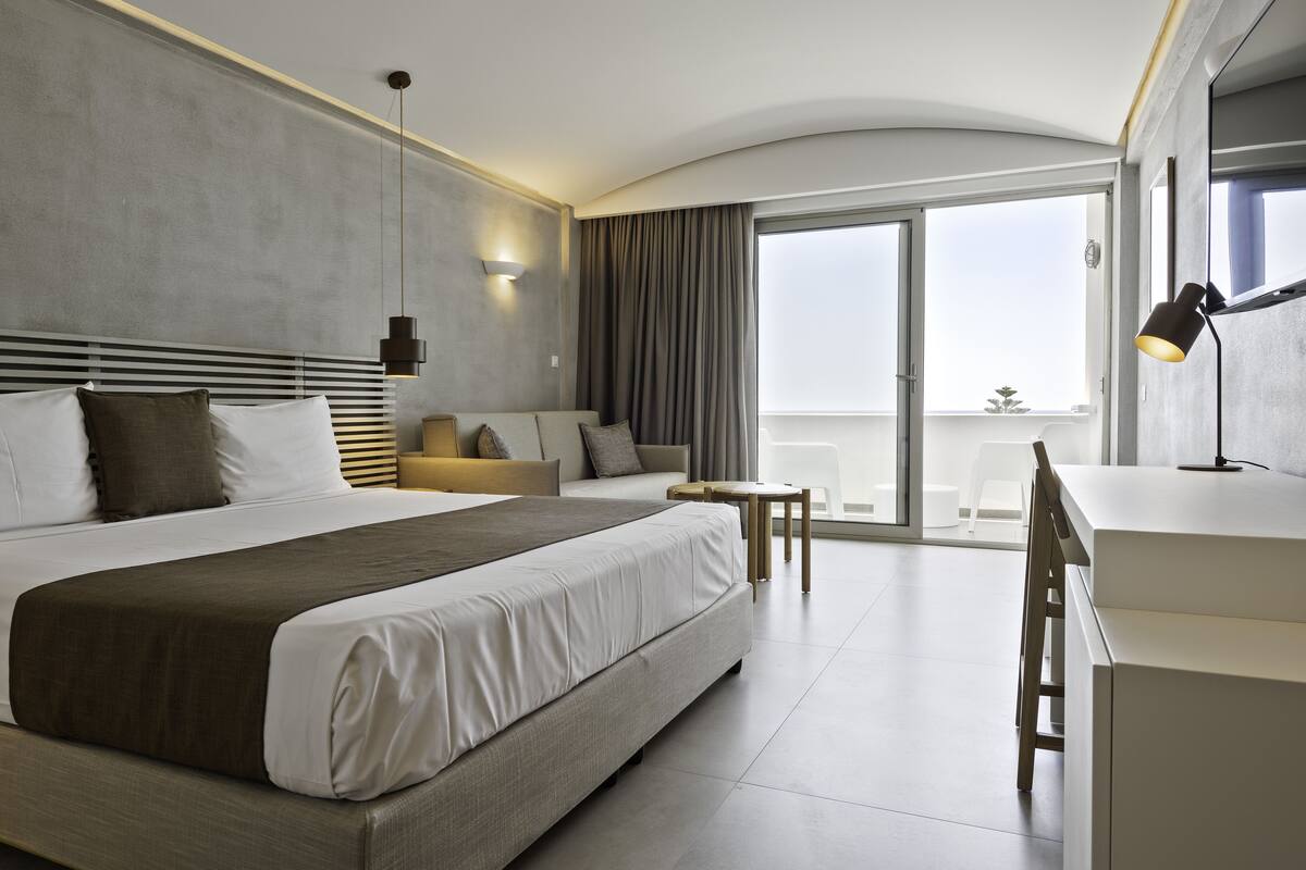 A modern hotel room with a large bed, a sofa, a desk, and a balcony with a view.