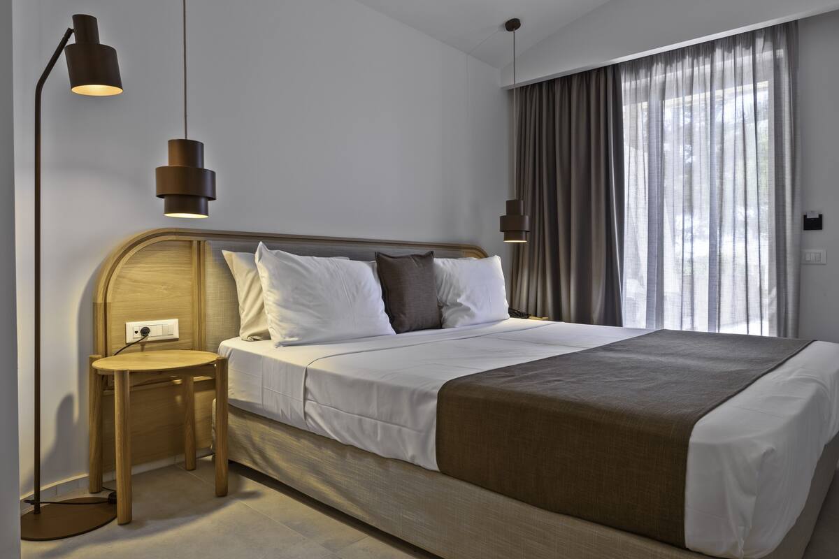 A modern hotel room with a large bed, a bedside table, a lamp, and a view of the outdoors through sheer curtains.