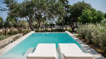 Una piscina rectangular con dos sillas reclinables blancas mirando al agua, rodeada de árboles y un borde de piedra.