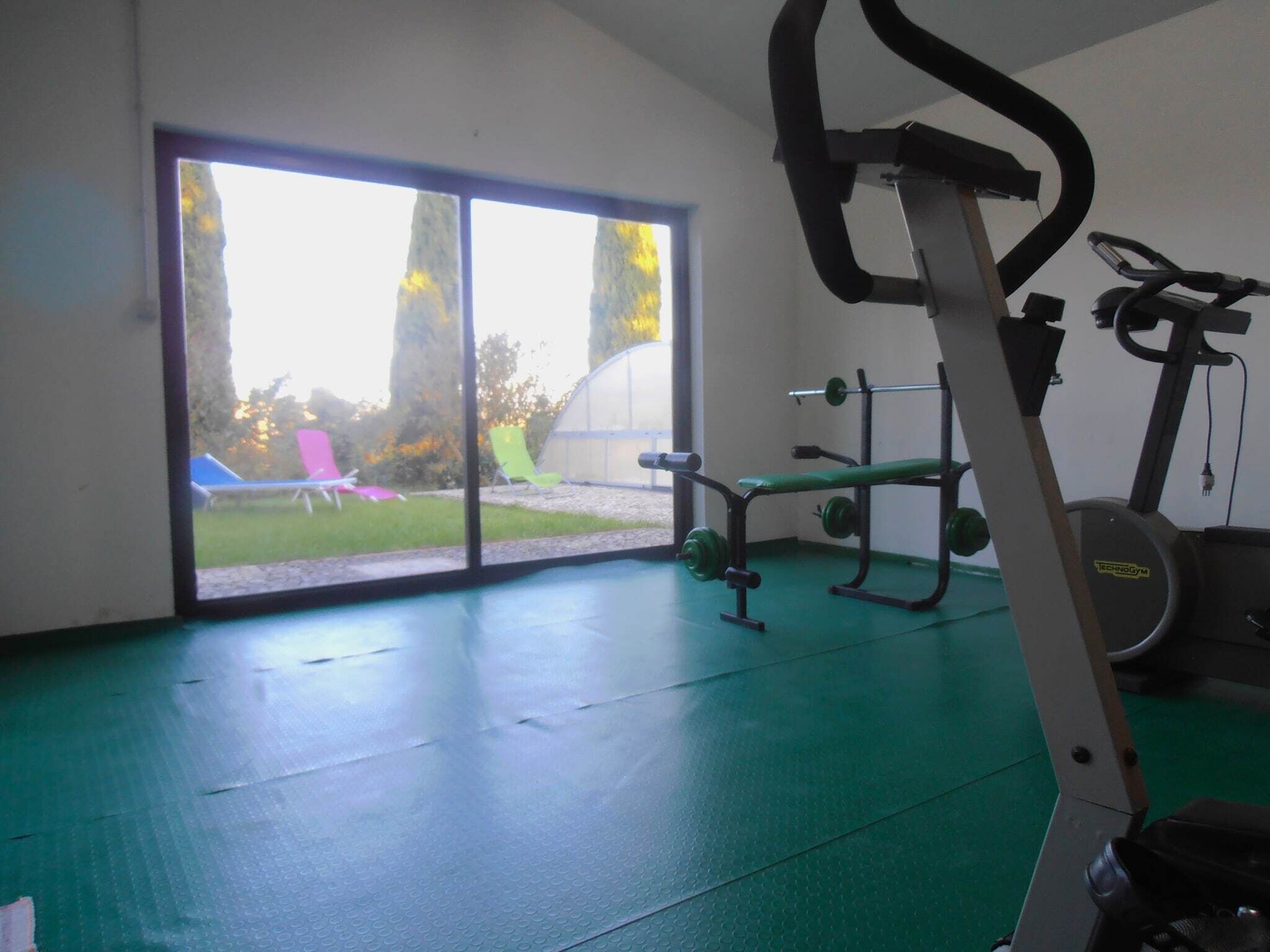 Une salle de sport équipée de matériel d’exercice, d’un banc et offrant une vue sur un espace extérieur aménagé avec des sièges et des arbres, le tout visible à travers une porte vitrée.