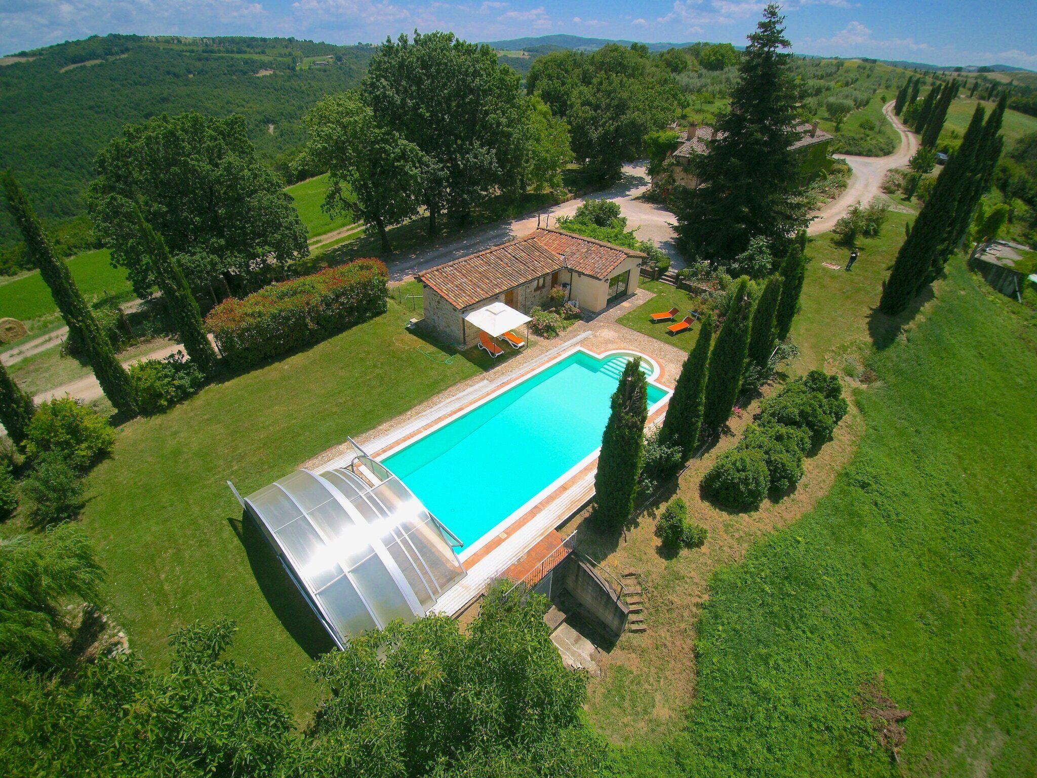 A large swimming pool with a glass-enclosed hot tub, surrounded by lush greenery and a small house.