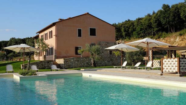 Una piscina con sillones de descanso y sombrillas, un muro de piedra y un edificio con ventanas y contraventanas.