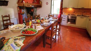 Un comedor con una larga mesa de madera dispuesta para una comida, una chimenea de ladrillo rojo y una cocina con armarios blancos y un horno de acero inoxidable.