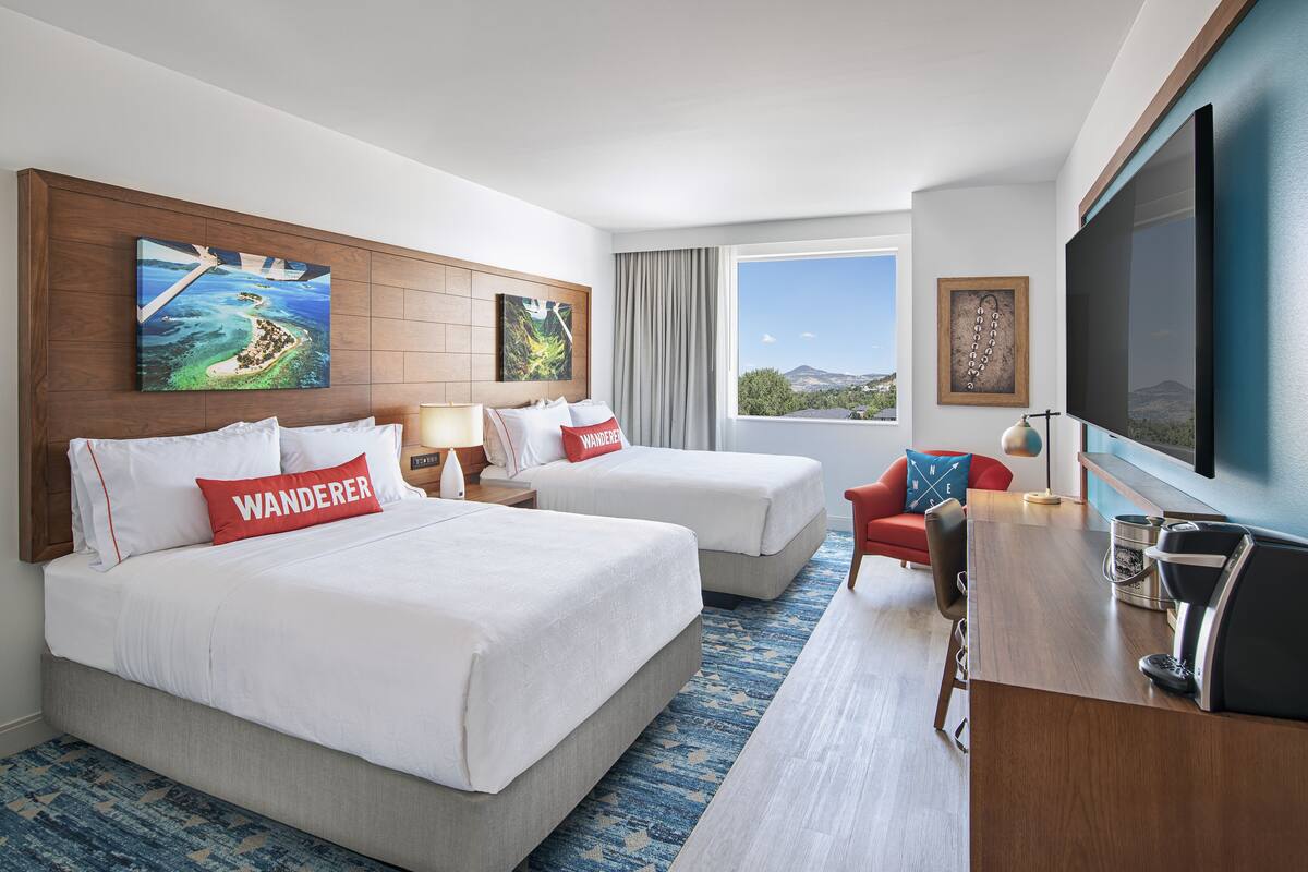A hotel room with two beds, a desk, a TV, and a window with a view of the mountains.