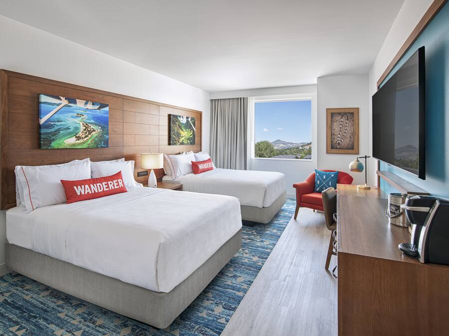 Habitación de hotel con dos camas, un escritorio, televisor y ventana con vistas a las montañas.