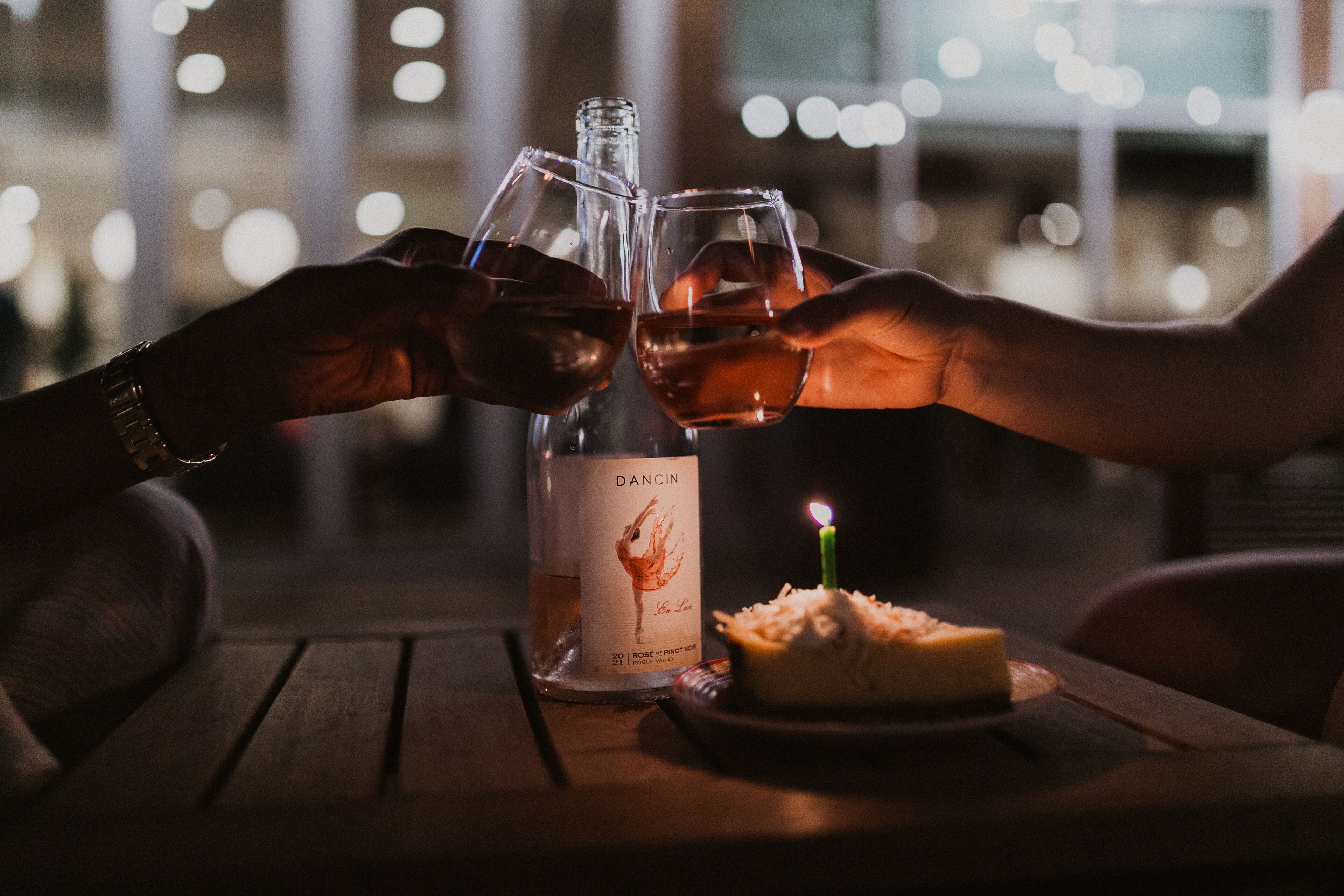 Dos manos chocando copas de vino junto a una botella de vino Dacinio y una vela sobre una mesa.