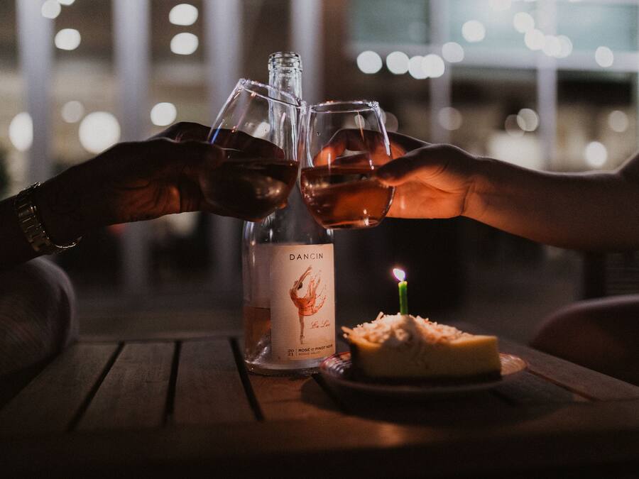 Two hands clinking wine glasses with a bottle of Dacinian wine and a candle on a table.