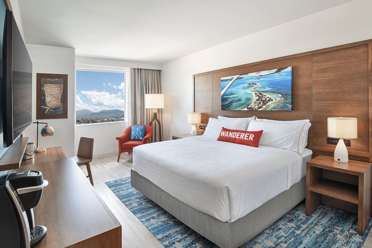 A hotel room with a large bed, a desk with a computer, a chair, a TV, and a window with a view of mountains and the sea.