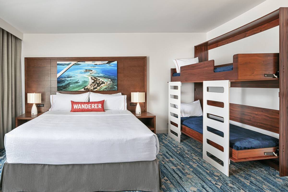 A hotel room with a large bed, a bunk bed, two bedside tables with lamps, a painting of a beach scene, and a 'Wanderer' pillow.