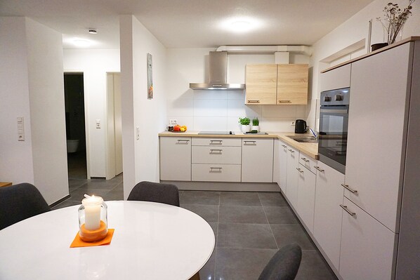A modern kitchen with white cabinets, a stainless steel range hood, and a built-in oven. There is a dining area with a round table and chairs, and a candle on the table.