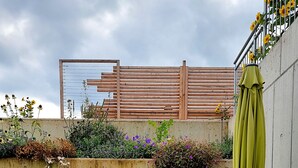 Une terrasse extérieure avec une table ronde en verre et des chaises en métal, entourée d’une variété de plantes et de fleurs.