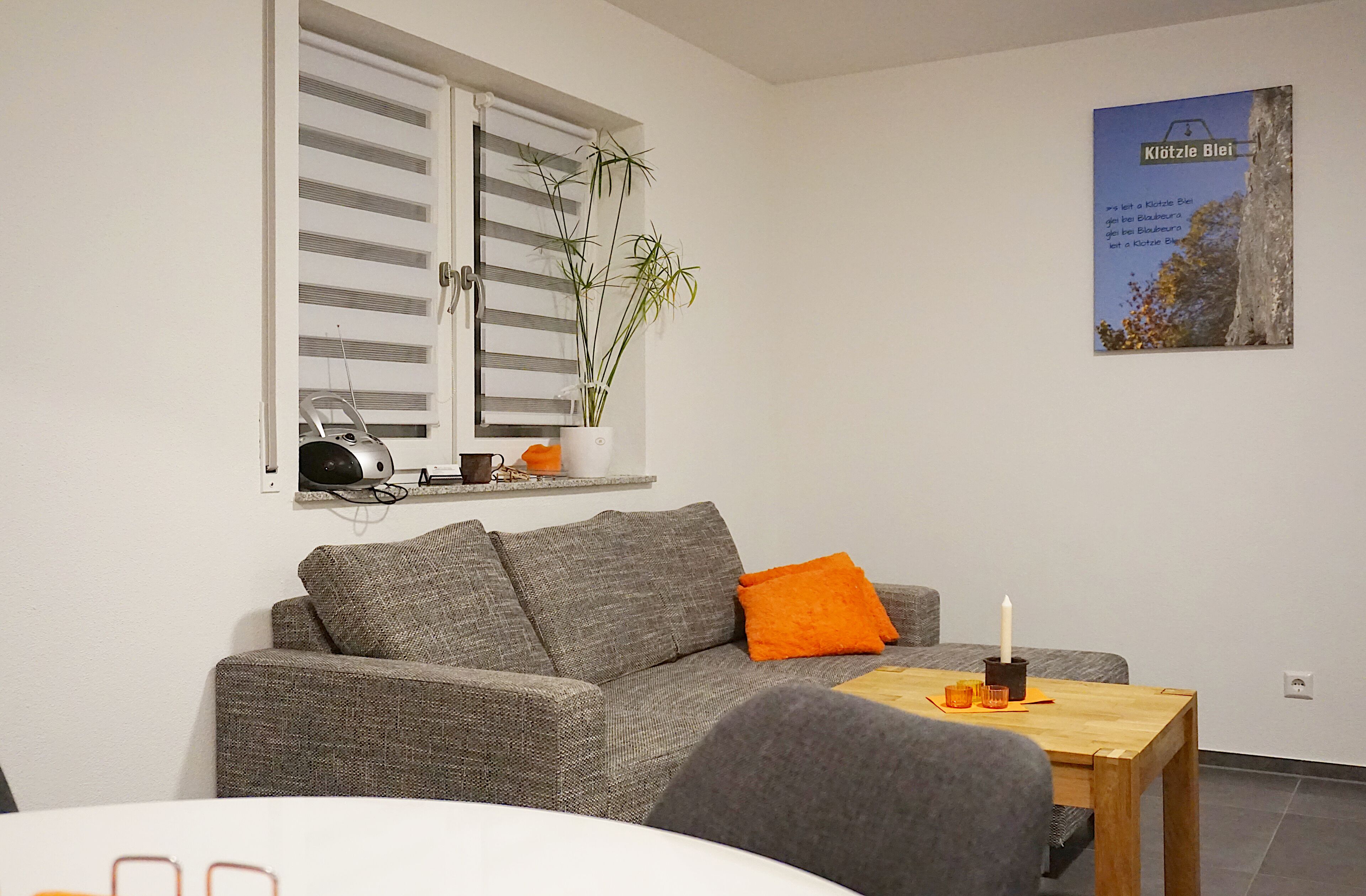 A living room with a grey sofa, a small table with a candle, and a window with blinds.