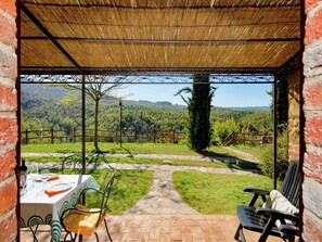 A covered outdoor seating area with a table set for two, a chair, and a view of a forested landscape. - Farmhouse in Città della Pieve with Pool (Città Della Pieve)