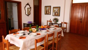 A dining room with a table set for six, surrounded by wooden chairs. There are two framed paintings on the wall, a wooden door, and a mirror.