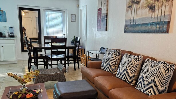 A living room with a brown sofa, a coffee table with a fruit basket, and a dining area with a table and chairs. -   5min drive to Universal studio (Orlando)