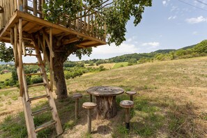 A rustic wooden structure with a ladder and a round table surrounded by stools, set in a grassy field with a tree and a distant view of hills. - Holiday Home in Assisi with Pool & Nature (Assisi)