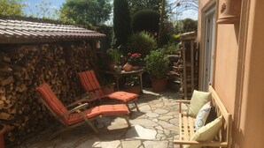 Un patio con suelo de piedra, dos sillones a rayas, una mesa con plantas en macetas y una estructura de madera con techo de tejas.