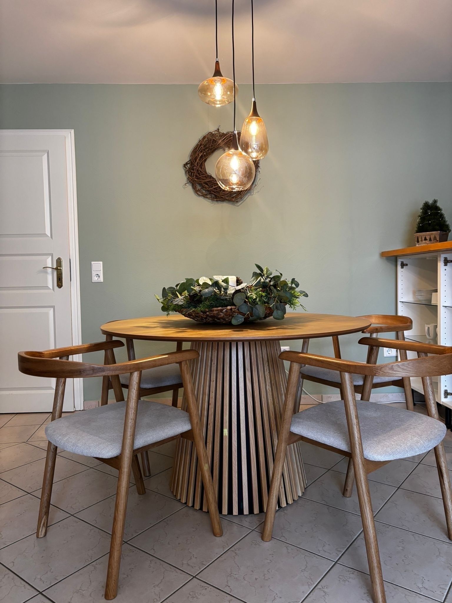 Un espace repas comprenant une table ronde en bois, six chaises en bois avec des coussins gris et trois lampes suspendues.