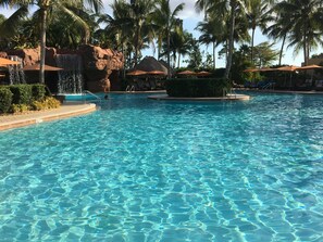 A clear pool with a rock formation and palm trees.