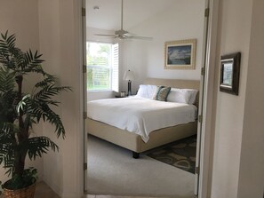 A bedroom with a bed, a ceiling fan, a window with blinds, a lamp, a framed picture on the wall, and a potted plant.