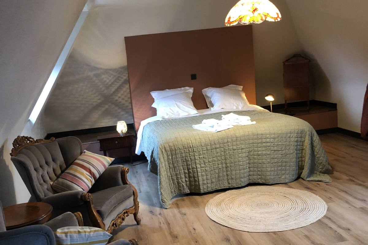 A bedroom with a sloped ceiling, a bed with a green quilt, a wooden nightstand, a blue armchair, and a round woven rug.