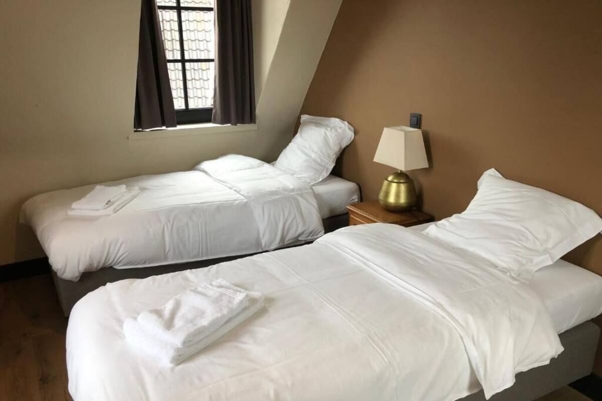 Two single beds with white linens, a bedside table with a lamp, and a window with curtains.